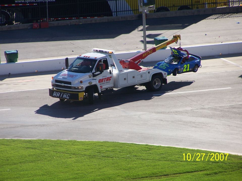 White tow truck lifting a blue race car with number 21, on a paved surface