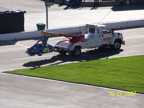 1979 Ford F-150 - A tow truck transports a blue race car with the number 21 on its side on a sunny day at a racetrack.