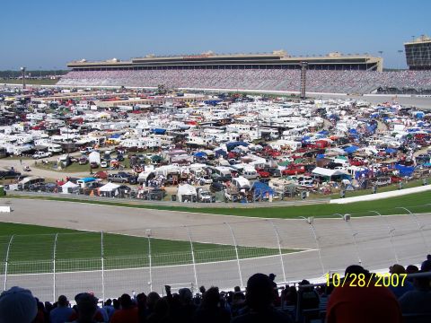 1979 Ford F-150 - A large crowd has gathered with their vehicles and RVs at a race track, with the grandstands visible in the distance.
