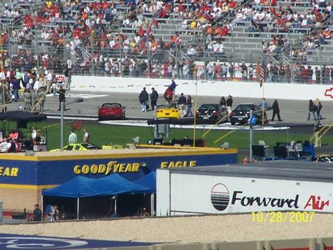 1979 Ford F-150 - A yellow sports car is elevated on a lift at a racetrack, with spectators in the stands and other vehicles on the track.