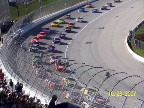 1979 Ford F-150 - A pack of stock cars races around a track, viewed from above with spectators in the stands.