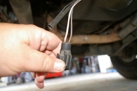 1986 Ford Bronco - A hand holds a disconnected wiring harness connector, revealing exposed wires beneath a vehicle.