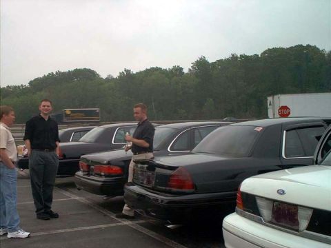 2000 Ford Crown Victoria - A group of people stand near several black Ford Crown Victoria sedans parked in an outdoor lot.