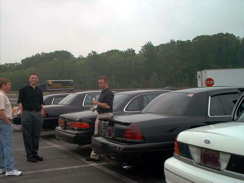 Several black Ford Crown Victorias parked in a lot with people standing around