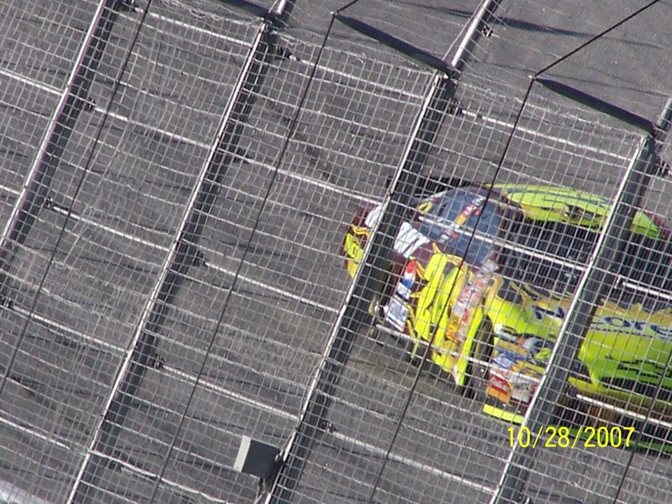 Two NASCAR race cars, one yellow and one green, on a track behind a fence. Date 10/28/2007 visible