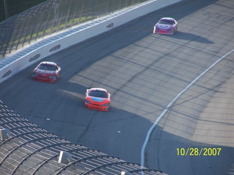 1979 Ford F-150 - Three stock cars compete on a paved race track under a bright sky, with shadows cast across the asphalt.