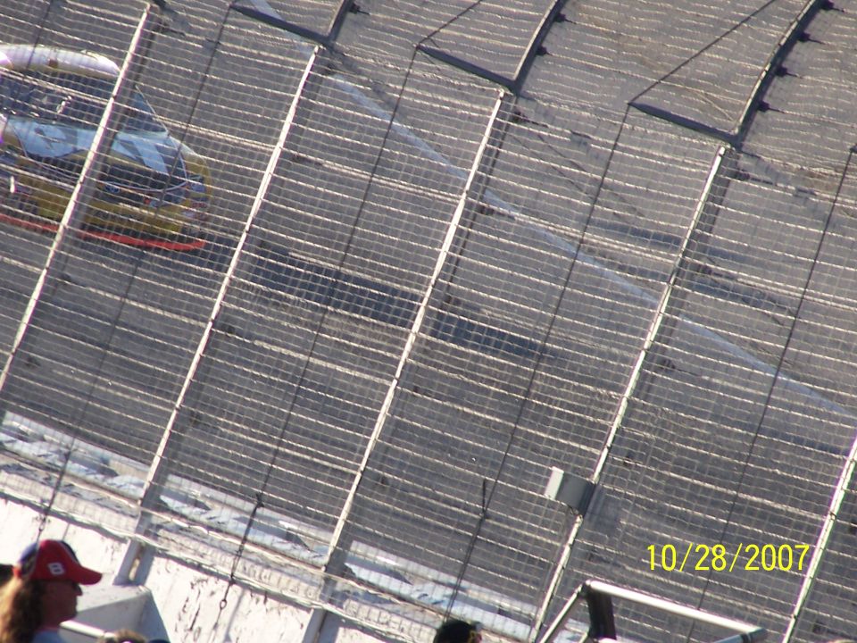 Race car with yellow and red livery on a track, seen from above through a fence