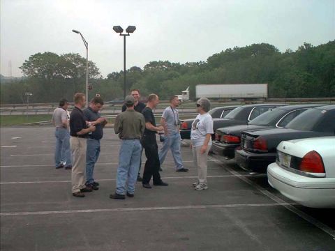 2000 Ford Crown Victoria - A group of people gather in a parking lot, with several sedans parked in a row behind them.
