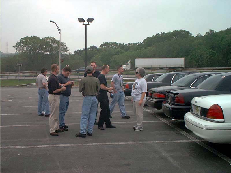 Several guys stand around in a parking lot, talking near a line of parked sedans