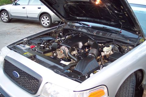 2001 Ford Crown Victoria - The engine bay of a 2001 Ford Crown Victoria is visible with the hood raised, and another car is parked behind it.