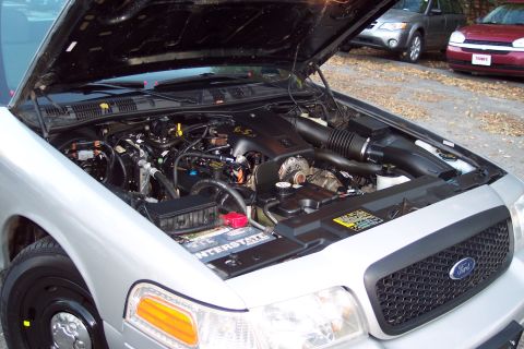 2001 Ford Crown Victoria - Engine bay of a silver Ford Crown Victoria, with a V8 engine prominently displayed. Other vehicles are parked in the background.