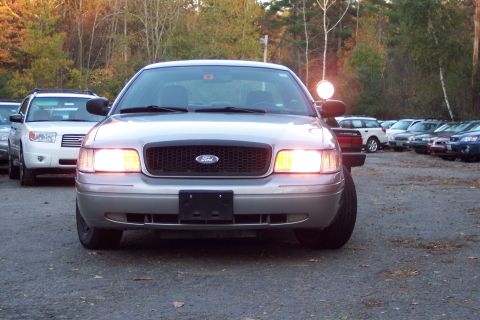 2001 Ford Crown Victoria - A silver 2001 Ford Crown Victoria is parked outdoors with its headlights illuminated, showing its front grille and Ford emblem.