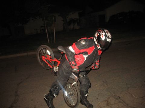 2007 Yamaha R1 - A person in full motorcycle gear performs a wheelie on a red bicycle under the cover of darkness.