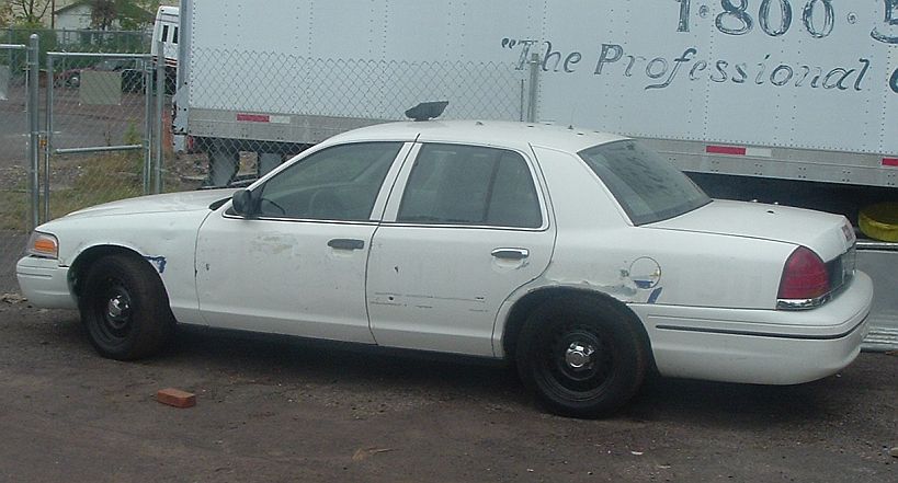 White Crown Victoria sedan with black steel wheels and spotlight, parked beside a trailer and chain-link fence