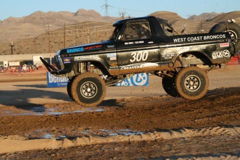 1990 Ford Bronco - black West Coast Broncos number 300 Ford-bodied trophy truck catches air over a desert dirt course lined with banners.