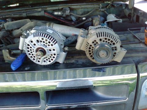 1995 Ford Bronco - Two alternators sitting side by side on the front core support of a dark green truck with the hood up, a clean new unit with a bare pulley…