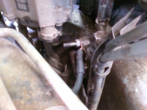 1995 Ford Bronco - Blurry low-light close-up down into a greasy engine bay, showing a cylinder-head area with a threaded stud and copper-washered fitting, a b…