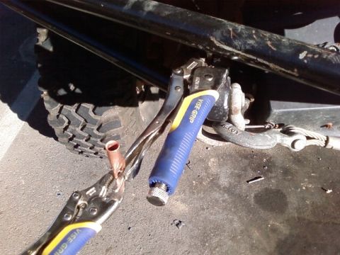 1995 Ford Bronco - Two blue-handled Vise-Grip locking pliers clamped onto a copper battery cable lug under a vehicle, with the black frame rail, a shackle mou…