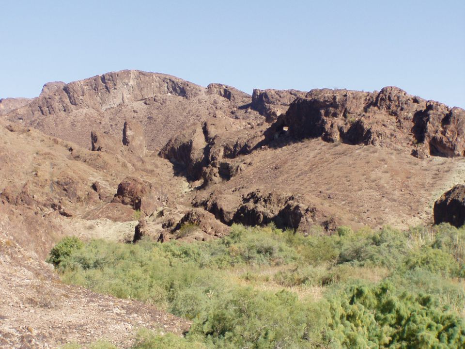 Rugged reddish-brown desert mountain with green brush at its base