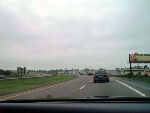 2000 Ford Crown Victoria - A dark sedan, likely a 2000 Ford Crown Victoria, travels on a highway under an overcast sky.