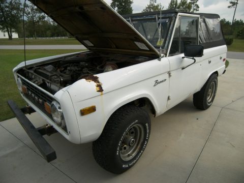1974 Ford Bronco - A white 1974 Ford Bronco is shown with its hood open, revealing the engine and some rust on the front fender.