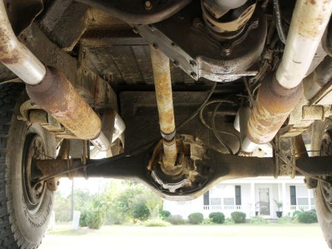 1974 Ford Bronco - The underside of a vehicle reveals its mechanical components, with a residential house visible beyond.