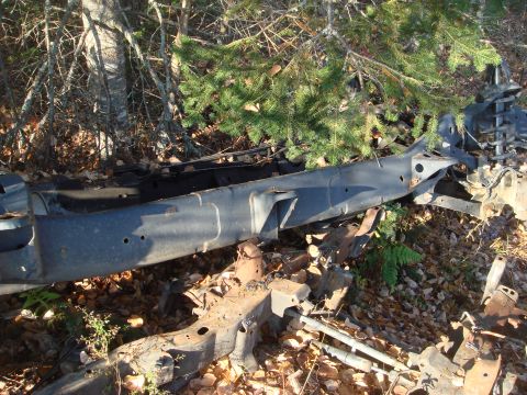 1996 Ford Bronco - The chassis and frame of a vehicle are visible, partially obscured by trees and fallen leaves in a wooded area.