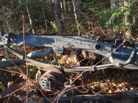 1996 Ford Bronco - Discarded vehicle frame and suspension components rest among fallen leaves in a wooded area.