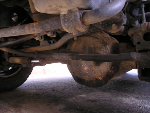 1989 Ford Bronco - The underside of a vehicle shows its front axle and suspension components, covered in dirt and rust.
