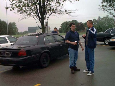 2000 Ford Crown Victoria - Two men converse next to a black 2000 Ford Crown Victoria in a parking lot.