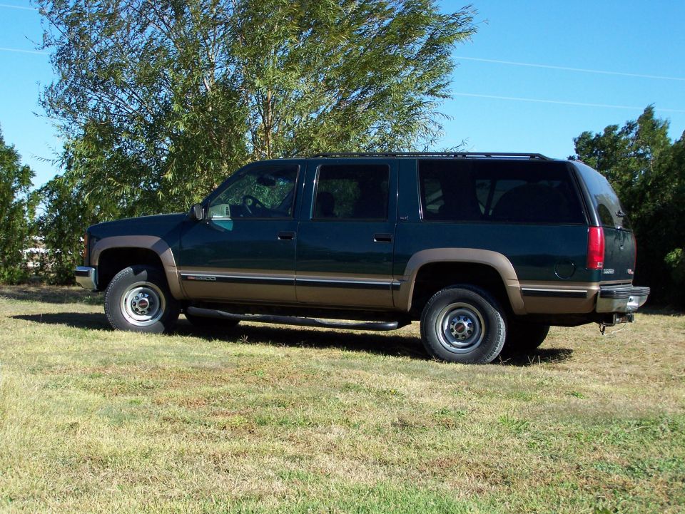 Green and brown two-tone SUV parked on grass with trees in background. Side view shows roof rack and chrome bumper