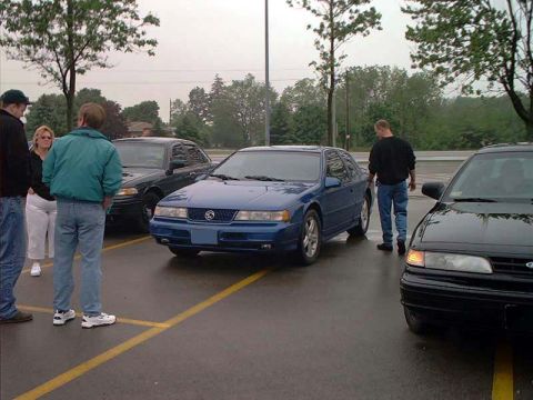 2000 Ford Crown Victoria - A blue sedan is parked in a lot, with other vehicles and people nearby.