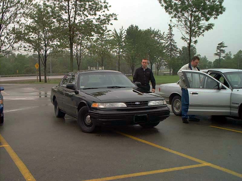 Black 2000 Ford Crown Victoria parked on wet asphalt, with a silver car to its right