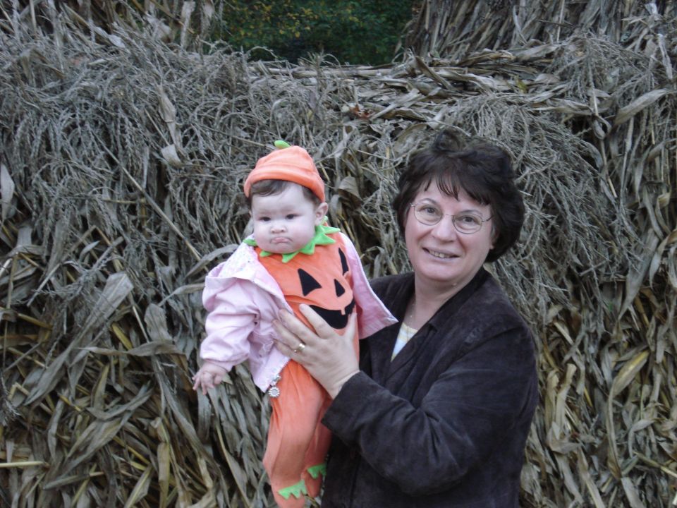 Woman holding baby dressed as a pumpkin in front of a corn stalk wall