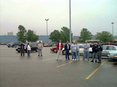 2000 Ford Crown Victoria - A group of people stand in a parking lot with multiple vehicles around them.