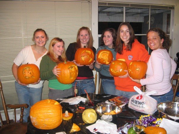 Group of six women holding carved pumpkins indoors, with a table of carving supplies in the foreground
