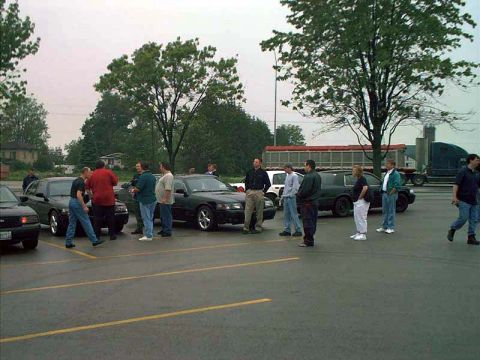 2000 Ford Crown Victoria - A group of men stand around several sedans in a parking lot on an overcast day.