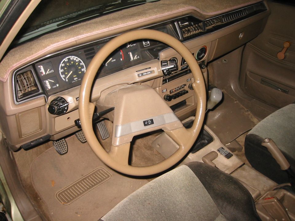Tan interior of a vehicle with beige steering wheel, analog gauges, dash cover, and manual shifter
