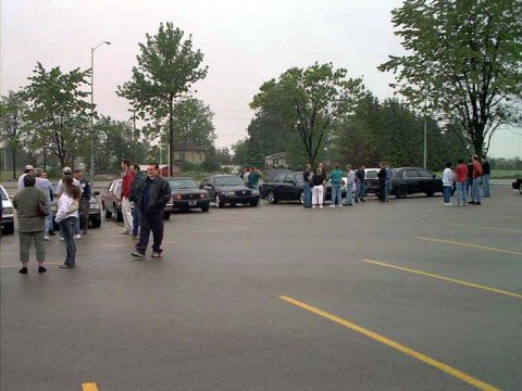 2000 Ford Crown Victoria - People gather among parked sedans at an outdoor car meet in a wet asphalt parking lot bordered by green trees.