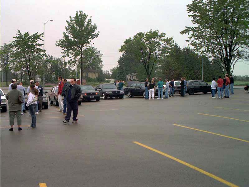 Crowd of people and parked cars at an outdoor car meet in a wet parking lot
