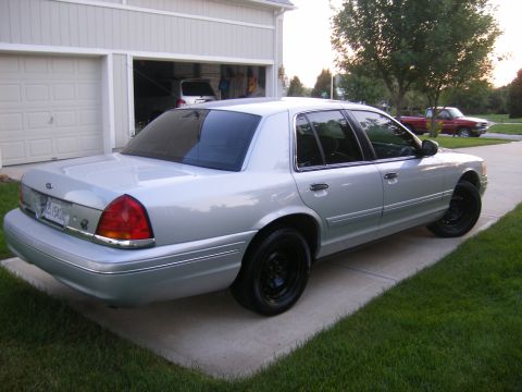 2001 Ford Crown Victoria - A silver 2001 Ford Crown Victoria is parked on a concrete driveway in front of a garage. A red truck is visible in the background.