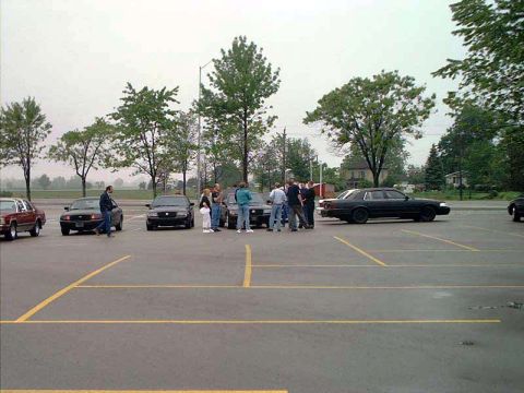 2000 Ford Crown Victoria - A group of people stand around several cars in a parking lot, including a black 2000 Ford Crown Victoria.