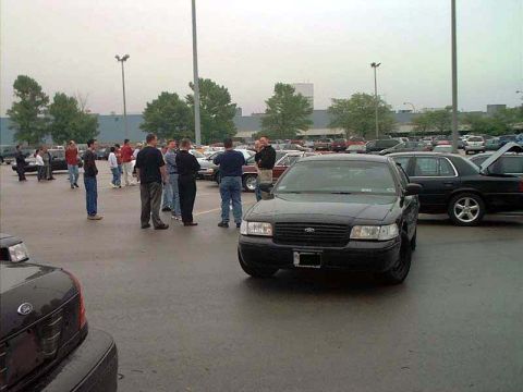 2000 Ford Crown Victoria - A group of people stand around a black 2000 Ford Crown Victoria in a parking lot.