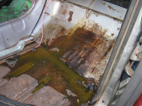 1979 Ford F-150 - Looking down into a stripped truck cab floor pan with carpet and seats removed, showing heavy surface rust and green coolant or fluid poole…