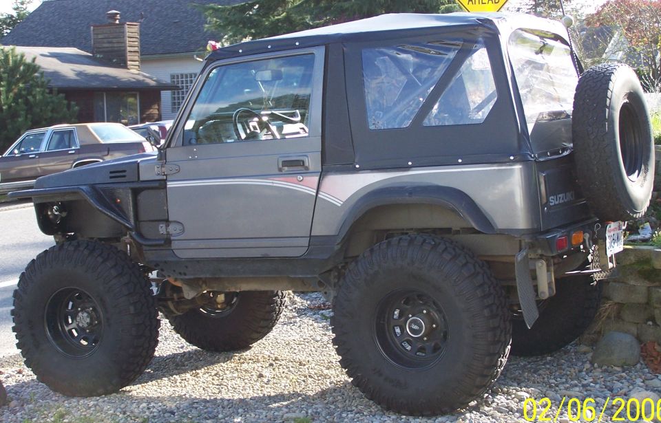 Lifted two-tone blue and gray Suzuki Samurai with soft top and big mud tires, driver side view