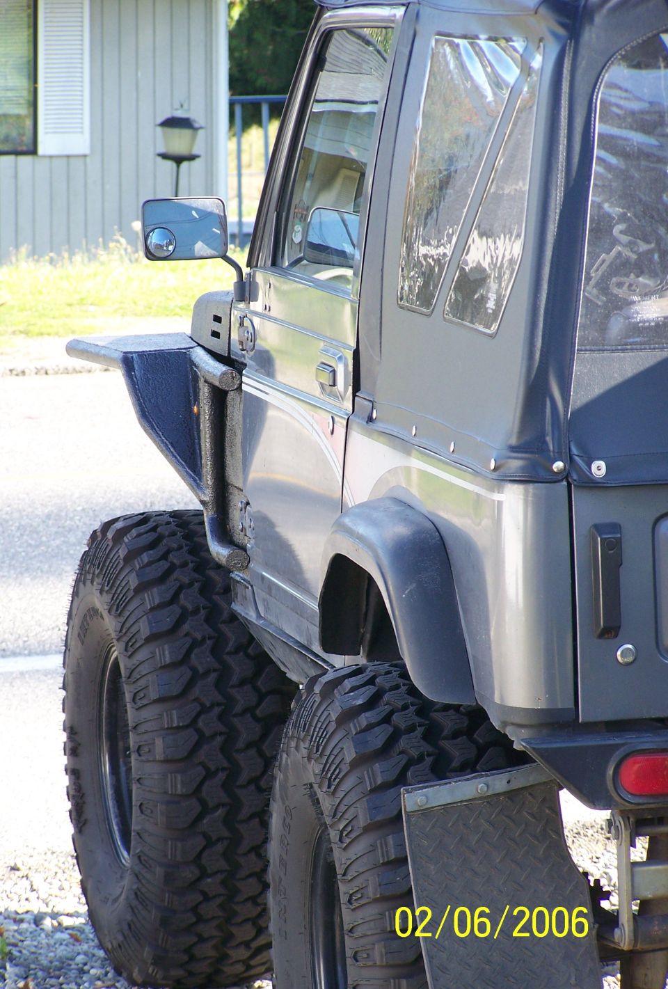 Side detail of lifted gray Suzuki Samurai: soft top, tube mirror mount, mud tires, diamond-plate flap