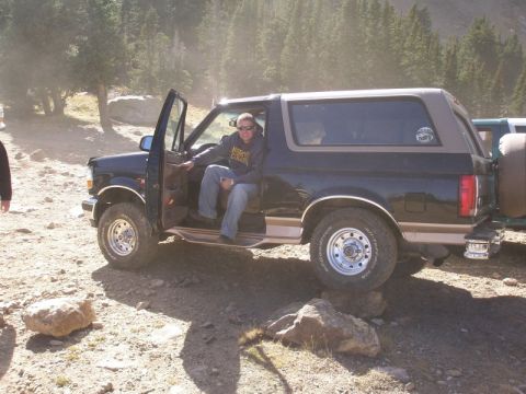 1996 Ford Bronco - A man sits in the driver's seat of a black and tan 1996 Ford Bronco, parked on a rocky trail with trees in the background.