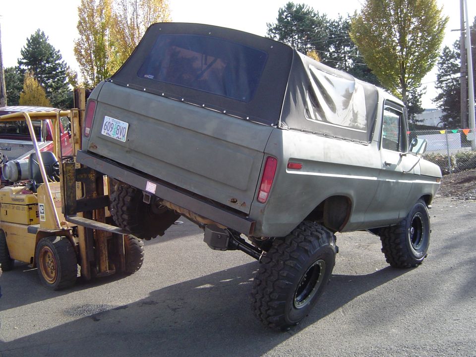 Olive green 1978 Ford Bronco lifted on large tires, being moved by a yellow forklift