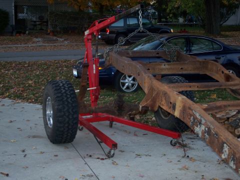 1979 Ford F-150 - A heavily rusted truck frame is supported by a red engine hoist, with a large tire nearby.