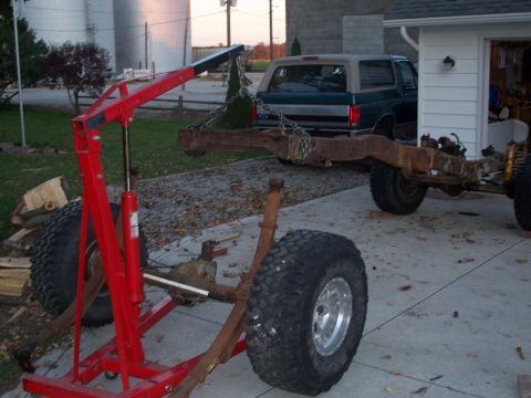 1979 Ford F-150 - A truck frame is suspended by a red engine hoist, with a green SUV visible behind it in a driveway.